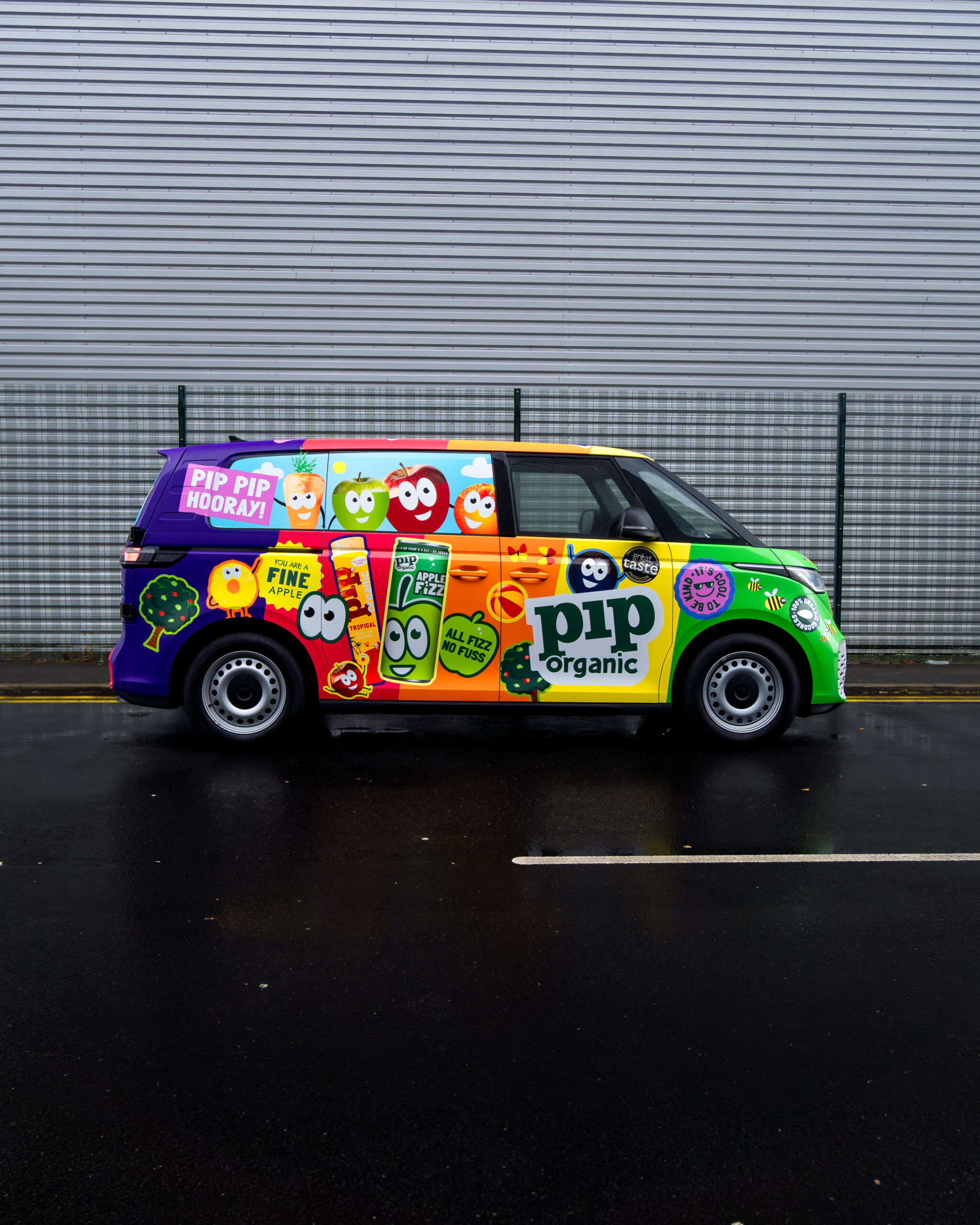 Colorful van with "Pip Organic" branding and cartoon fruit characters parked on a wet street, with a metal fence and a gray building in the background.