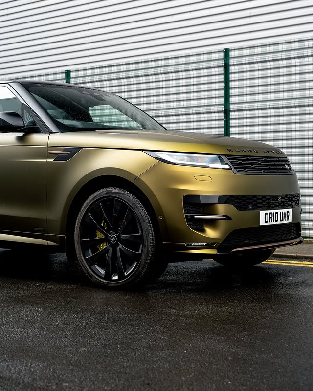 A greenish-gold SUV with a personalized license plate is parked on wet ground in front of a striped industrial wall.