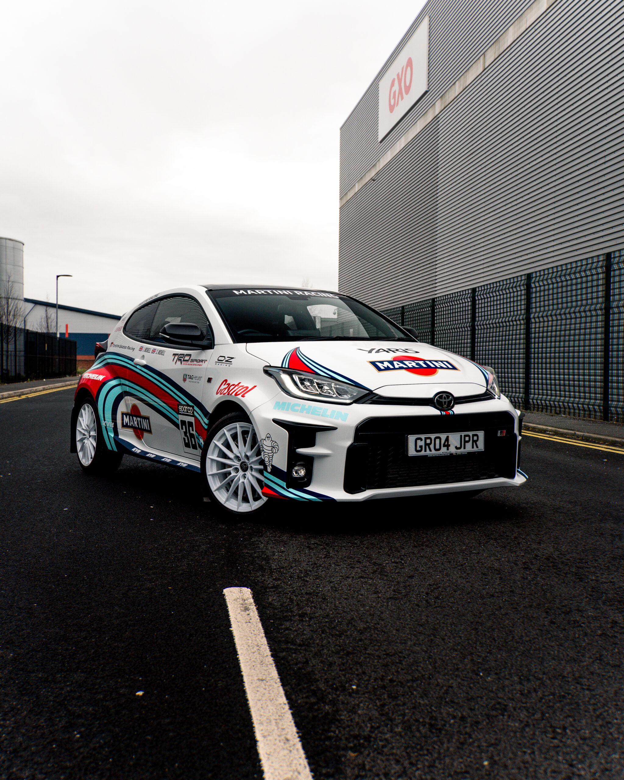 Rally car with racing decals, including Martini and Michelin, parked on a paved road near industrial buildings.