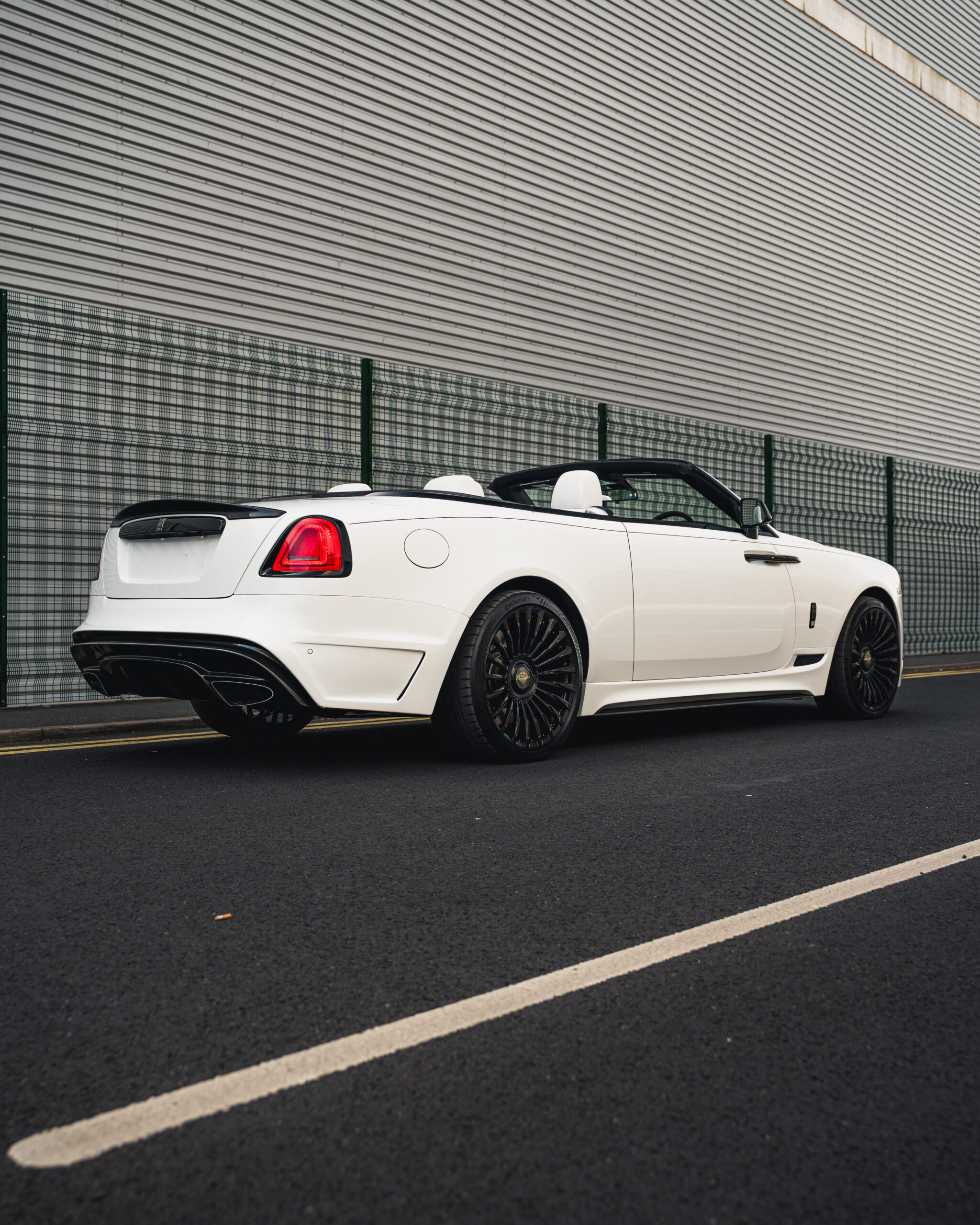 A white convertible luxury car is parked on a road next to a metal fence and a corrugated building wall.