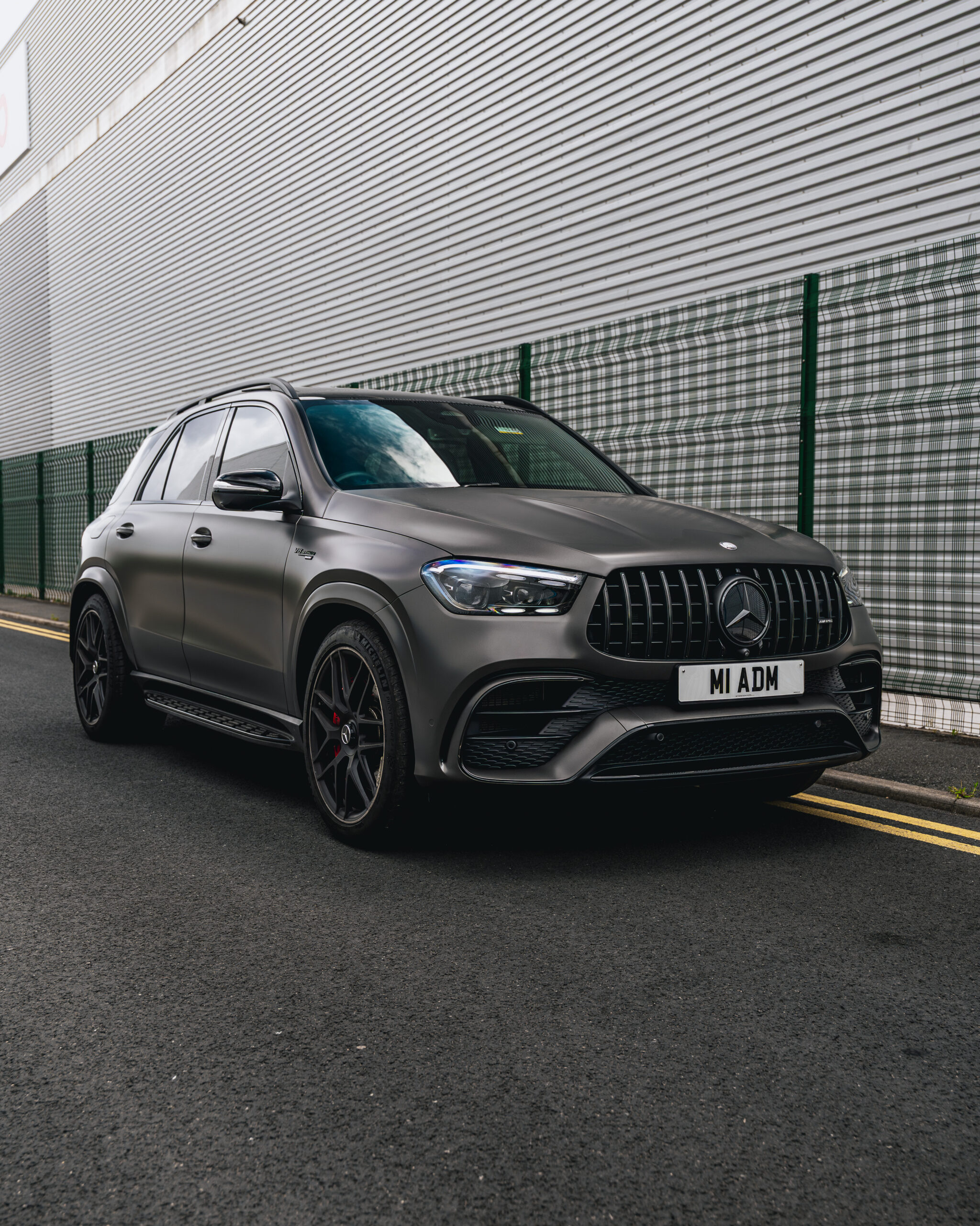 A matte grey Mercedes-Benz SUV is parked on a street next to a metal fence and a corrugated building, displaying a license plate reading "M1 ADM.