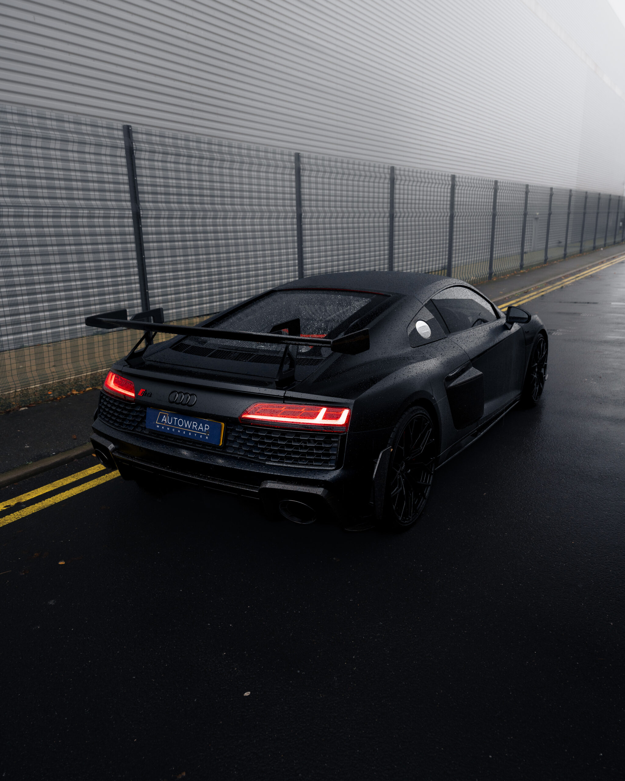 A black Audi R8 sports car with a rear spoiler is parked on a wet, empty street near a metal fence and industrial building on a foggy day.