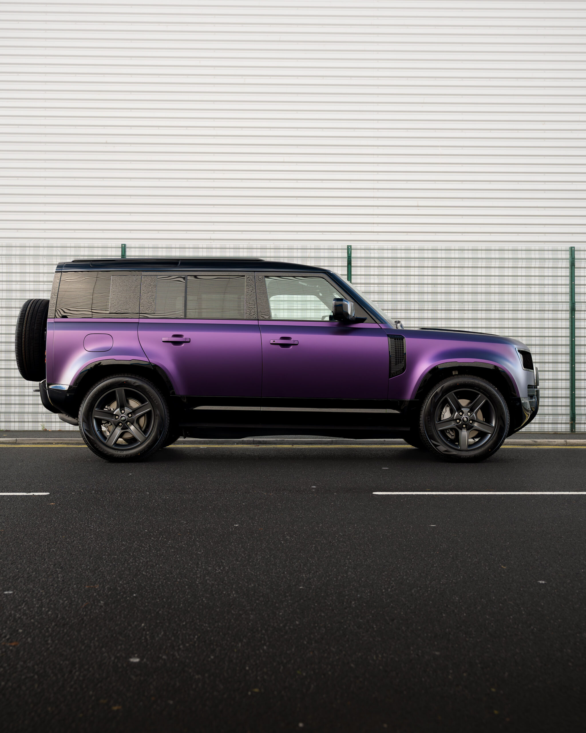 A purple SUV with black accents is parked on a street in front of a white metal wall with a green fence.