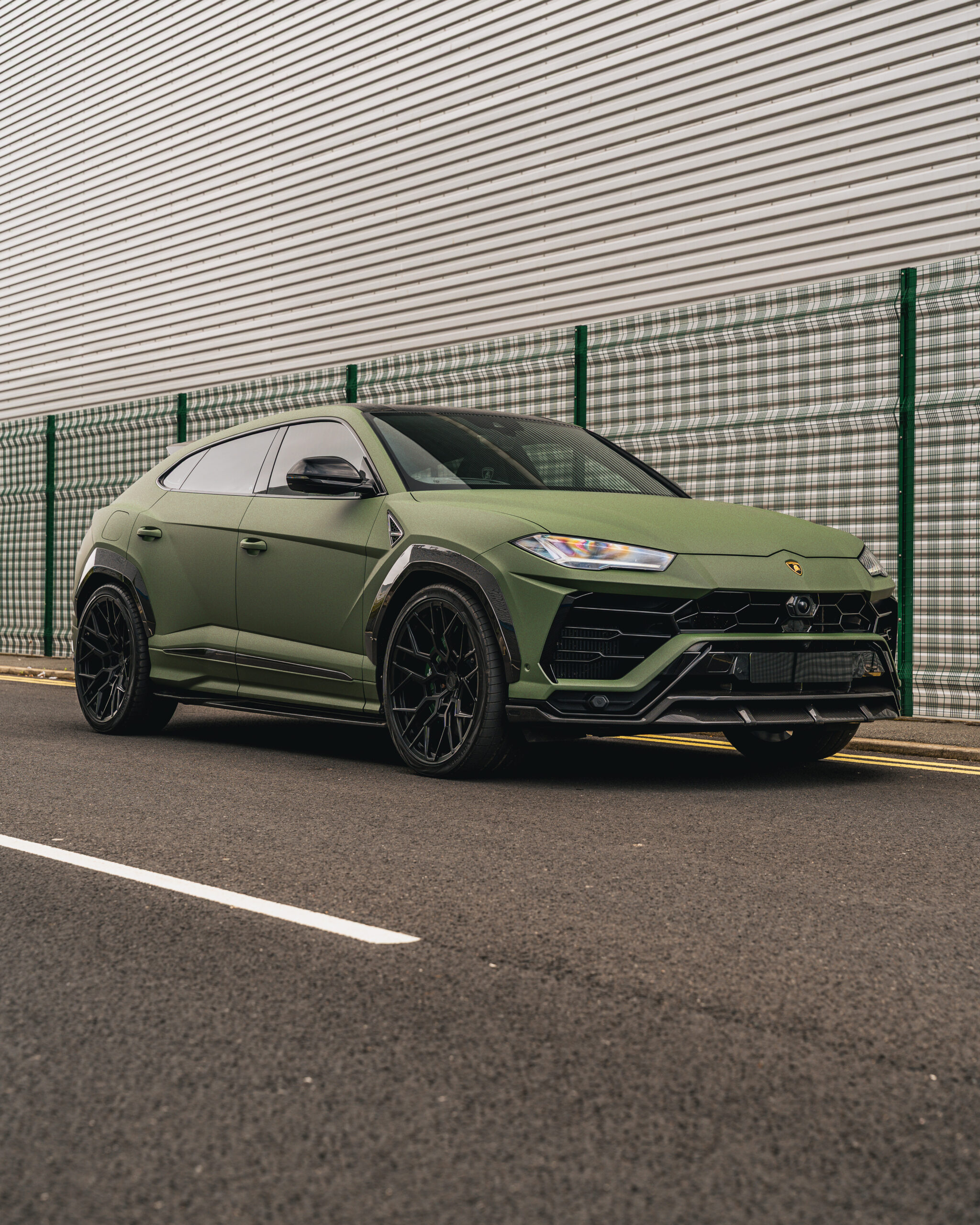 A matte green Lamborghini Urus SUV is parked on an asphalt road beside a large, corrugated metal building with a green fence.