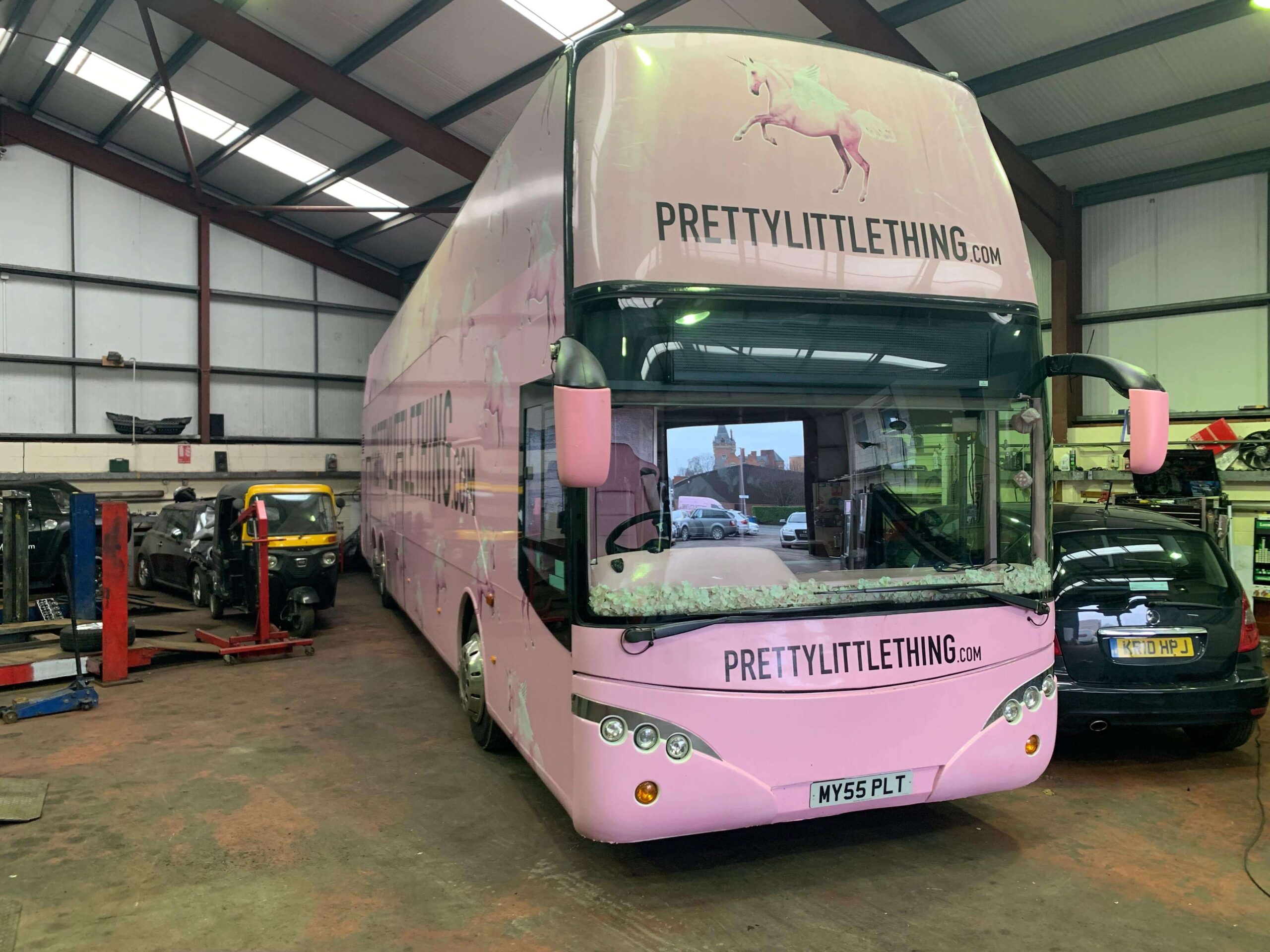 A pink double-decker bus with "PRETTYLITTLETHING.COM" branding is parked inside a garage with other vehicles nearby.