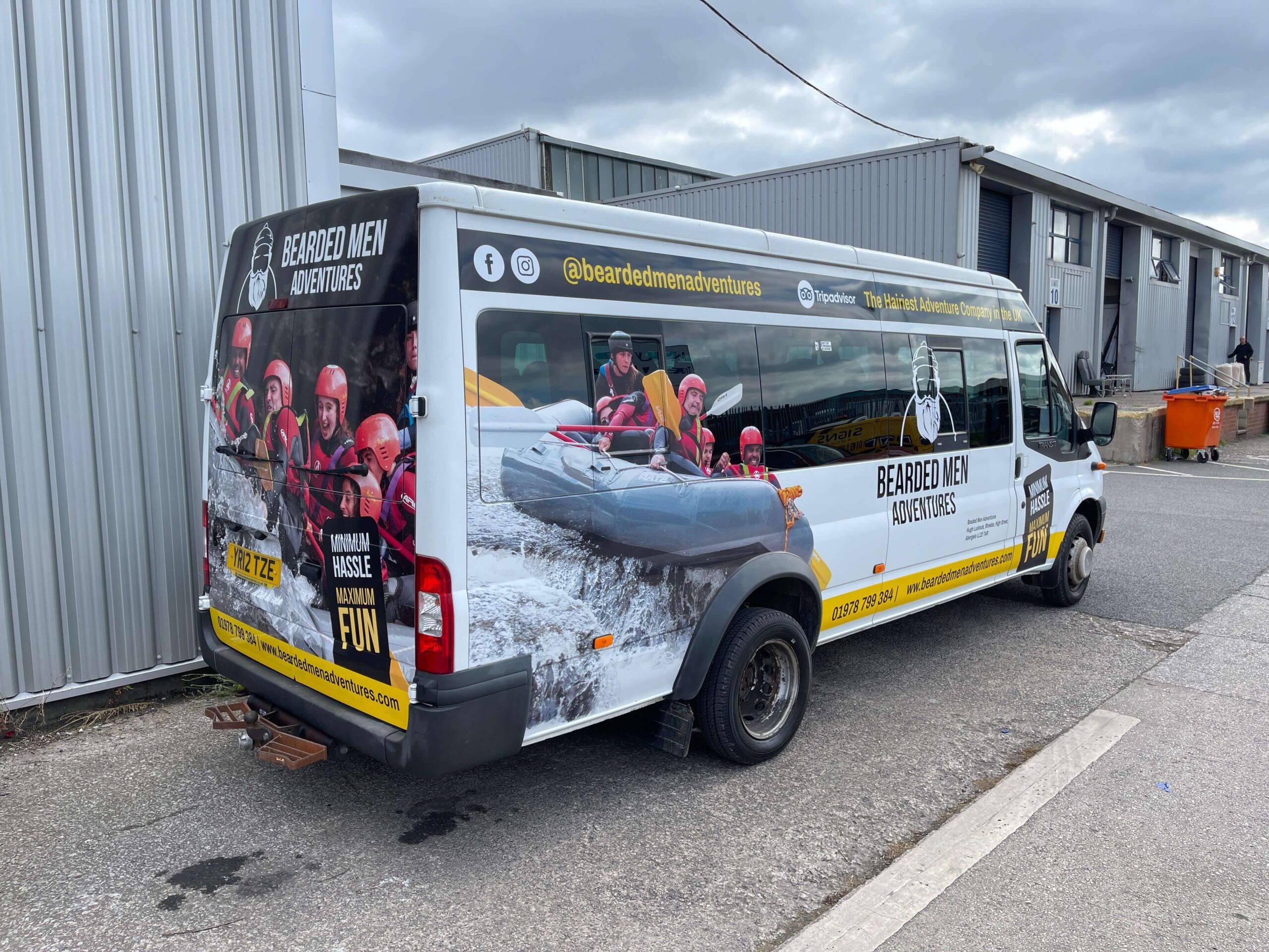 A white van branded with "Bearded Men Adventures" features images of people rafting, social media handles, and company contact details, parked beside an industrial building.