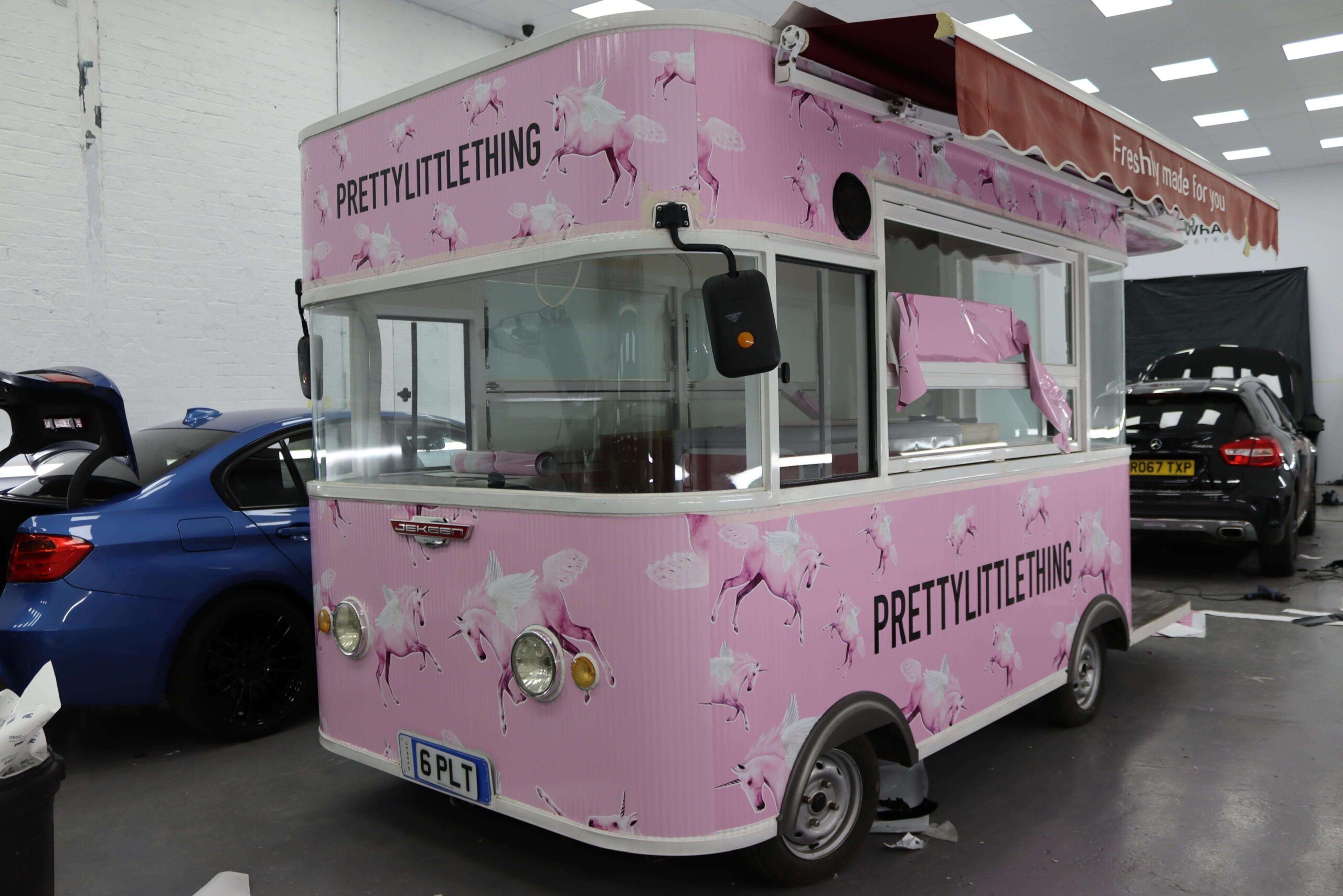 A small pink food truck with "PRETTYLITTLETHING" branding and unicorn graphics, parked indoors near several cars.