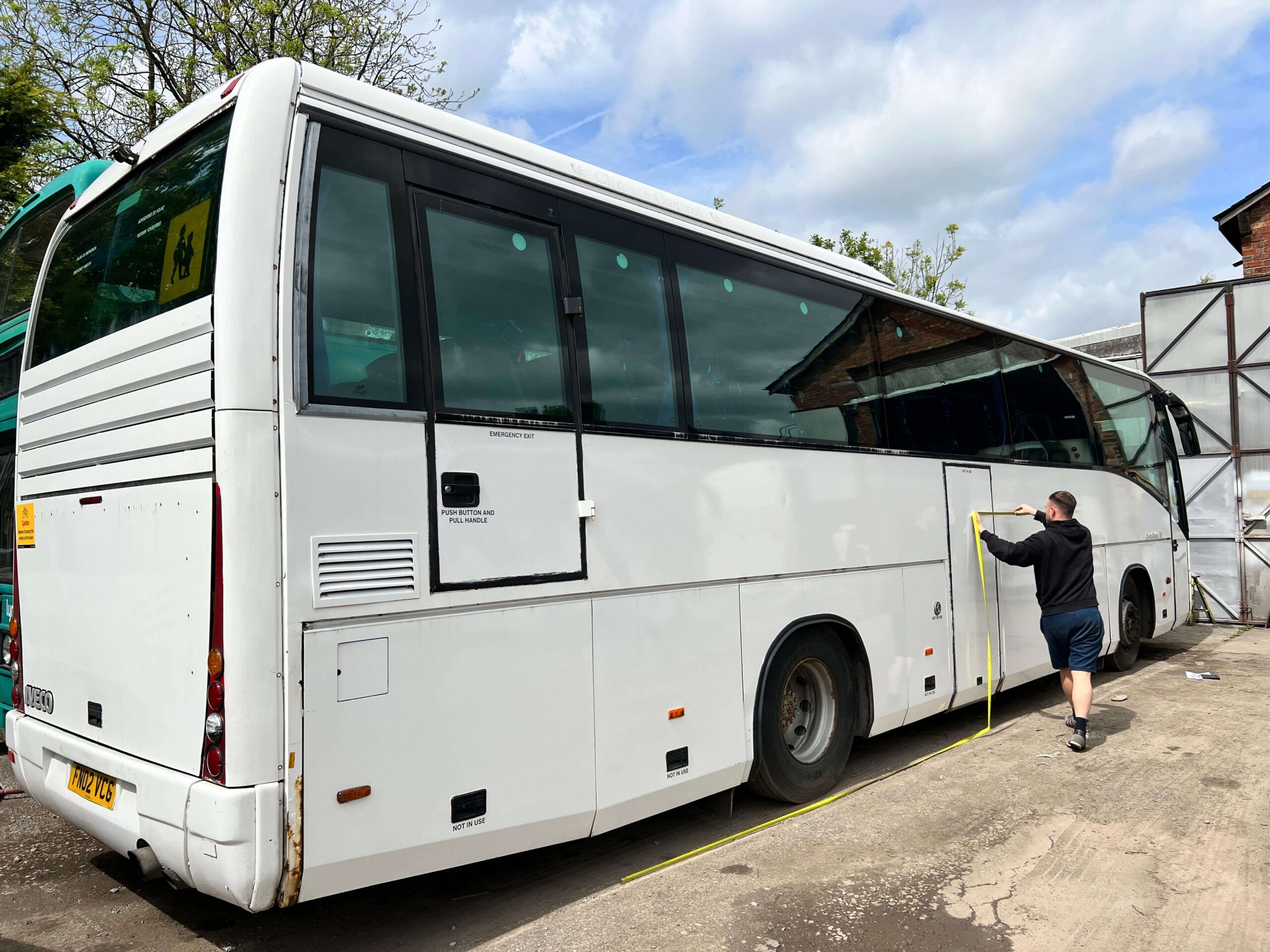 A person measures the length of a white coach bus parked outside near a building on a cloudy day.