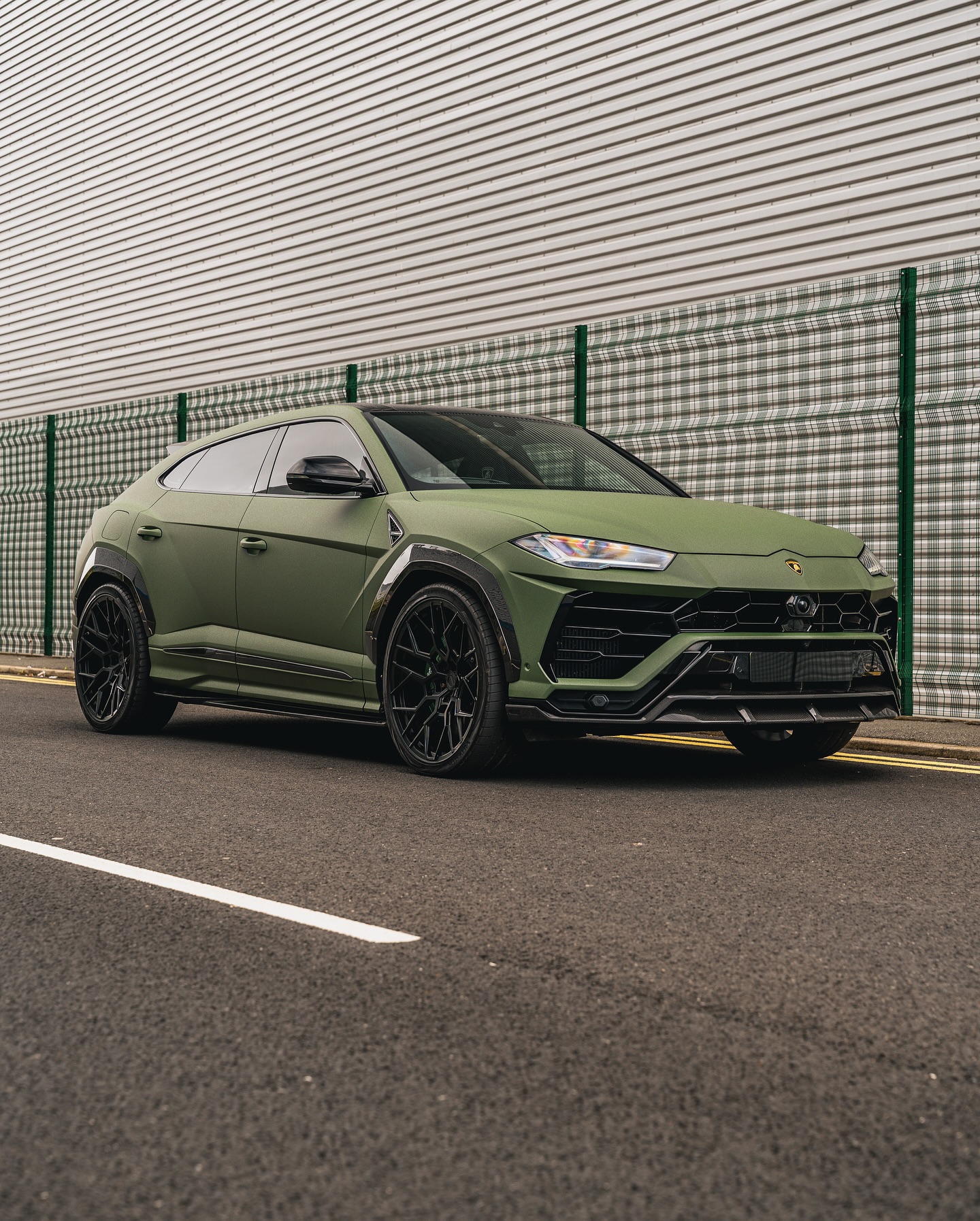 A matte green Lamborghini Urus SUV is parked on the side of a street next to a corrugated metal wall and a fence.