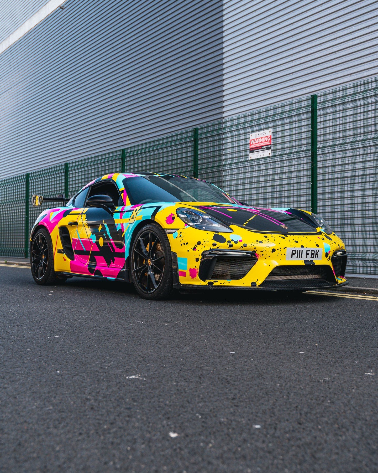 A sports car with a bright, multicolored graffiti-style wrap is parked on the street beside a metal fence and industrial building.