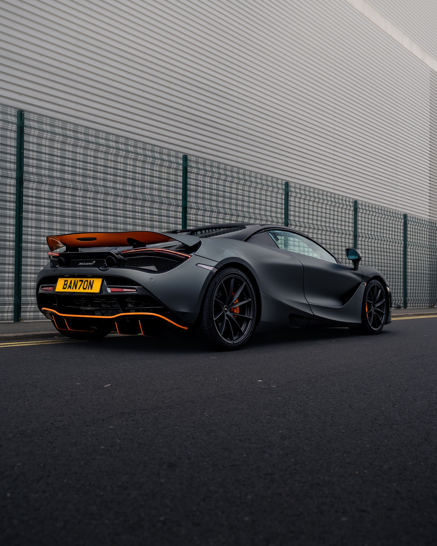 A black sports car with orange accents is parked on a street beside a tall, striped building and a metal fence.