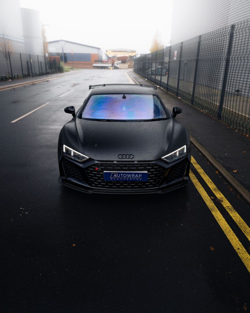 A matte black Audi sports car with rain droplets on the hood is parked on a wet street between industrial buildings.