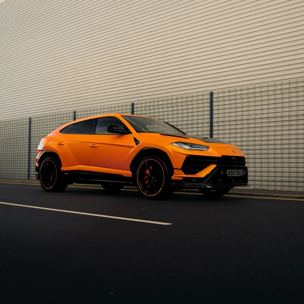 An orange luxury SUV is parked on a street beside a metal fence and a large industrial building with horizontal siding.