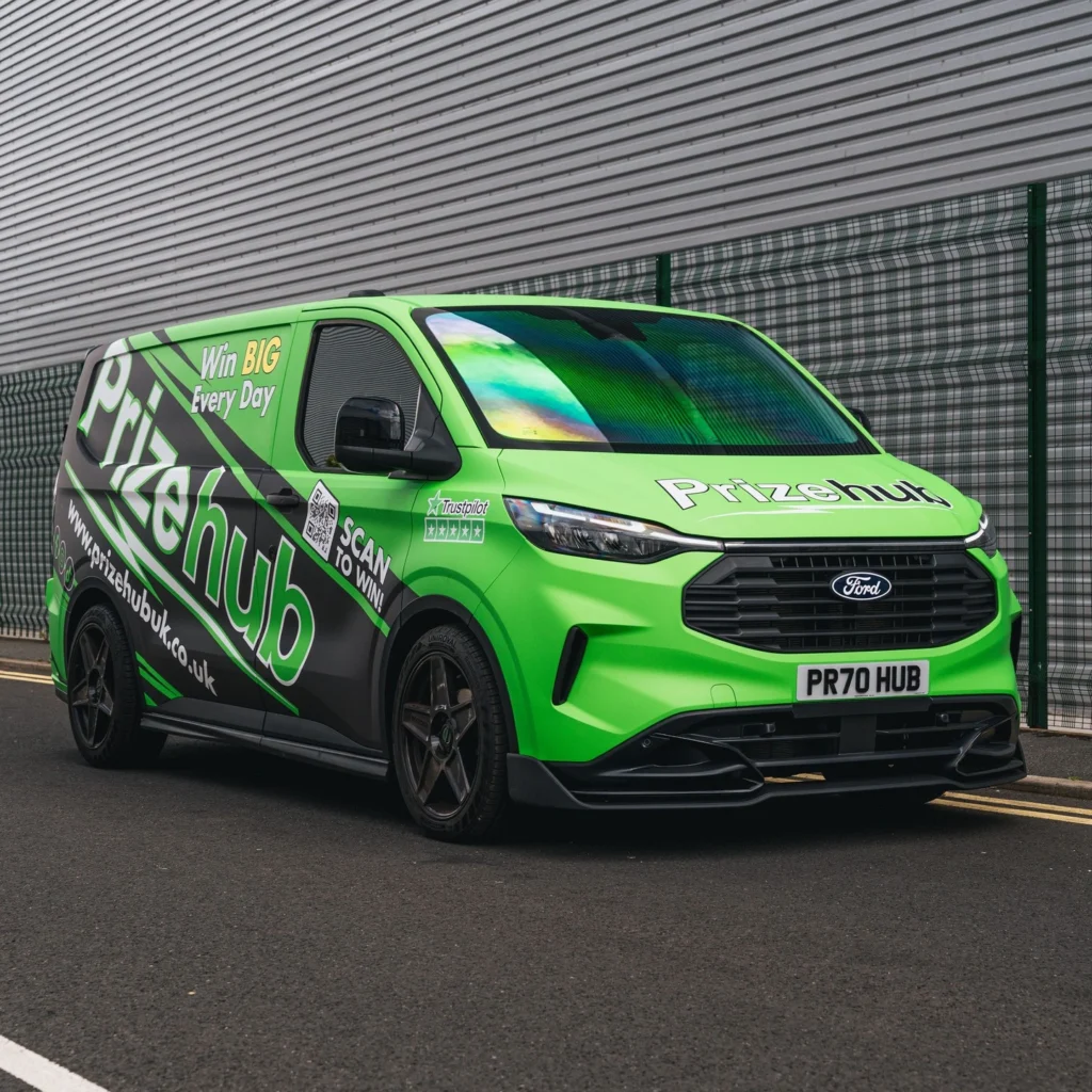 A green and black Ford van with "Prizehub" branding and promotional text is parked on a street beside a wire fence and metal building.