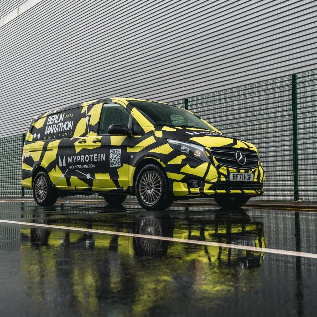 A black and yellow MyProtein-branded van with "Berlin Marathon" signage is parked on a wet street beside a metal fence and corrugated building.
