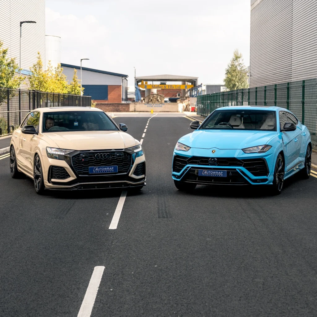 A beige Audi and a blue Lamborghini are parked side by side on a street between industrial buildings.