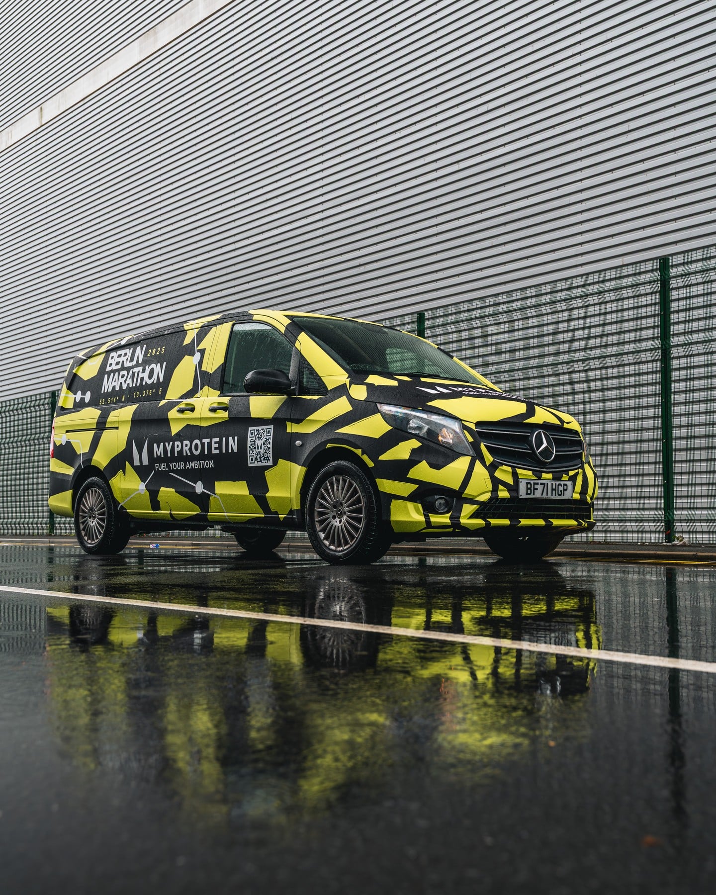 A yellow and black camouflaged Mercedes-Benz van with "Berlin Marathon" and "MyProtein" branding is parked on a wet street near a corrugated metal wall and fence.