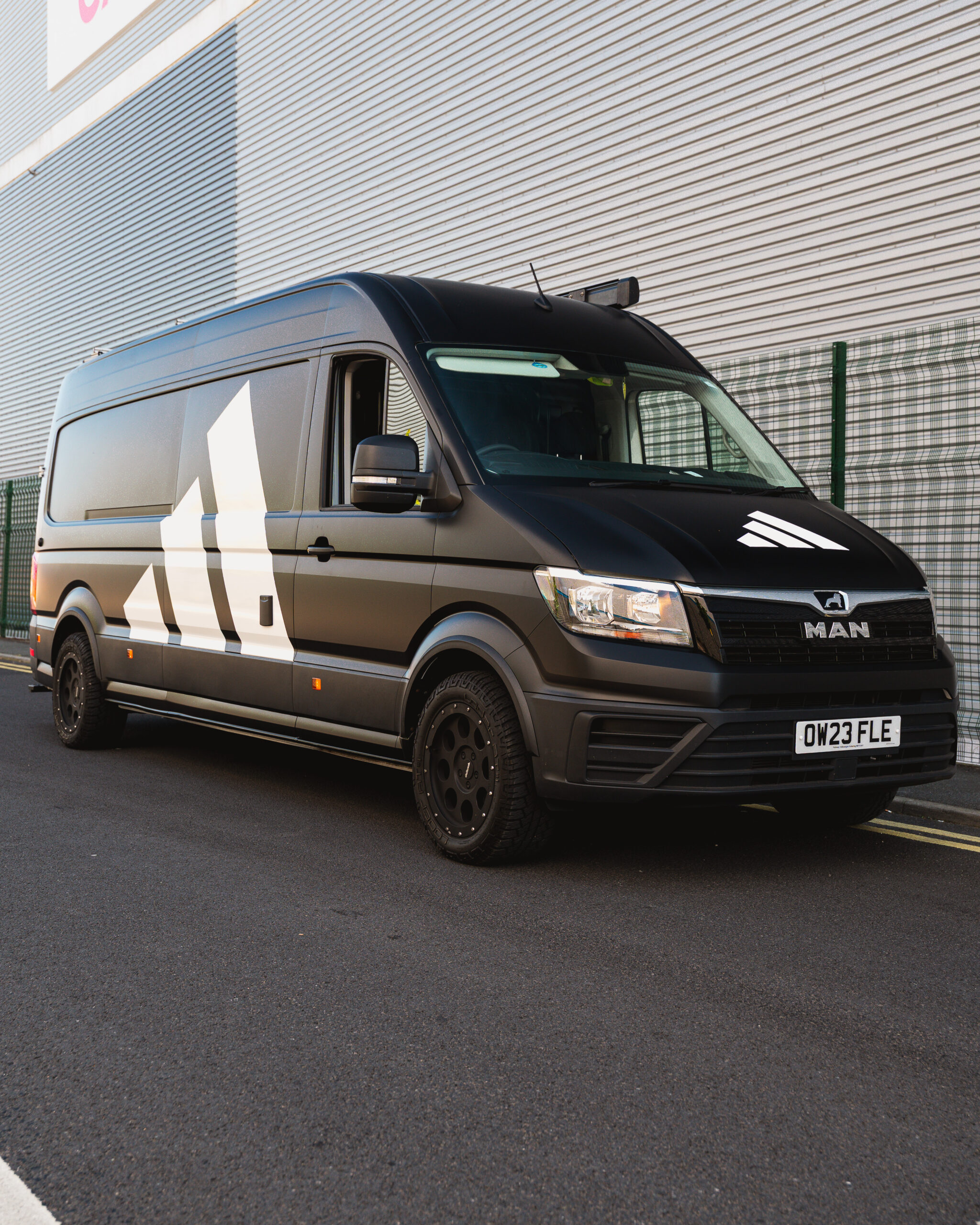 A black van with the Adidas logo on its side is parked on a street next to a metal fence and a corrugated building.