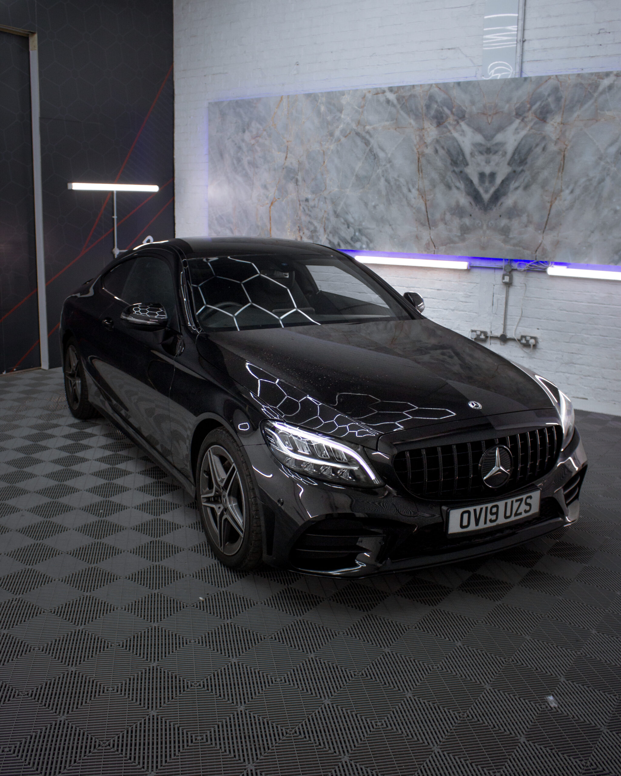 A black Mercedes-Benz coupe with license plate OV19 UZS is parked indoors on a patterned floor, with geometric lights reflecting on the car's surface.