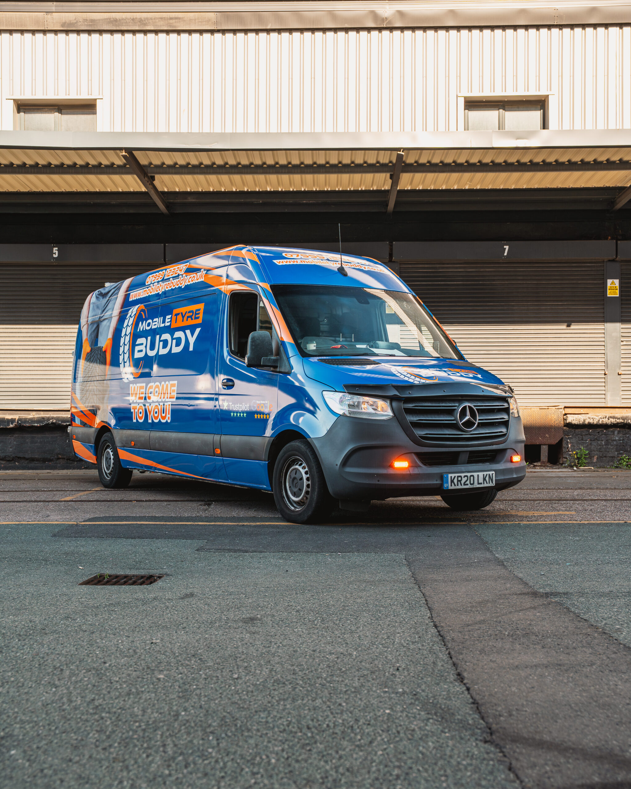 A blue and orange Mobile Tyre Buddy van is parked on a street in front of a warehouse with closed shutters.