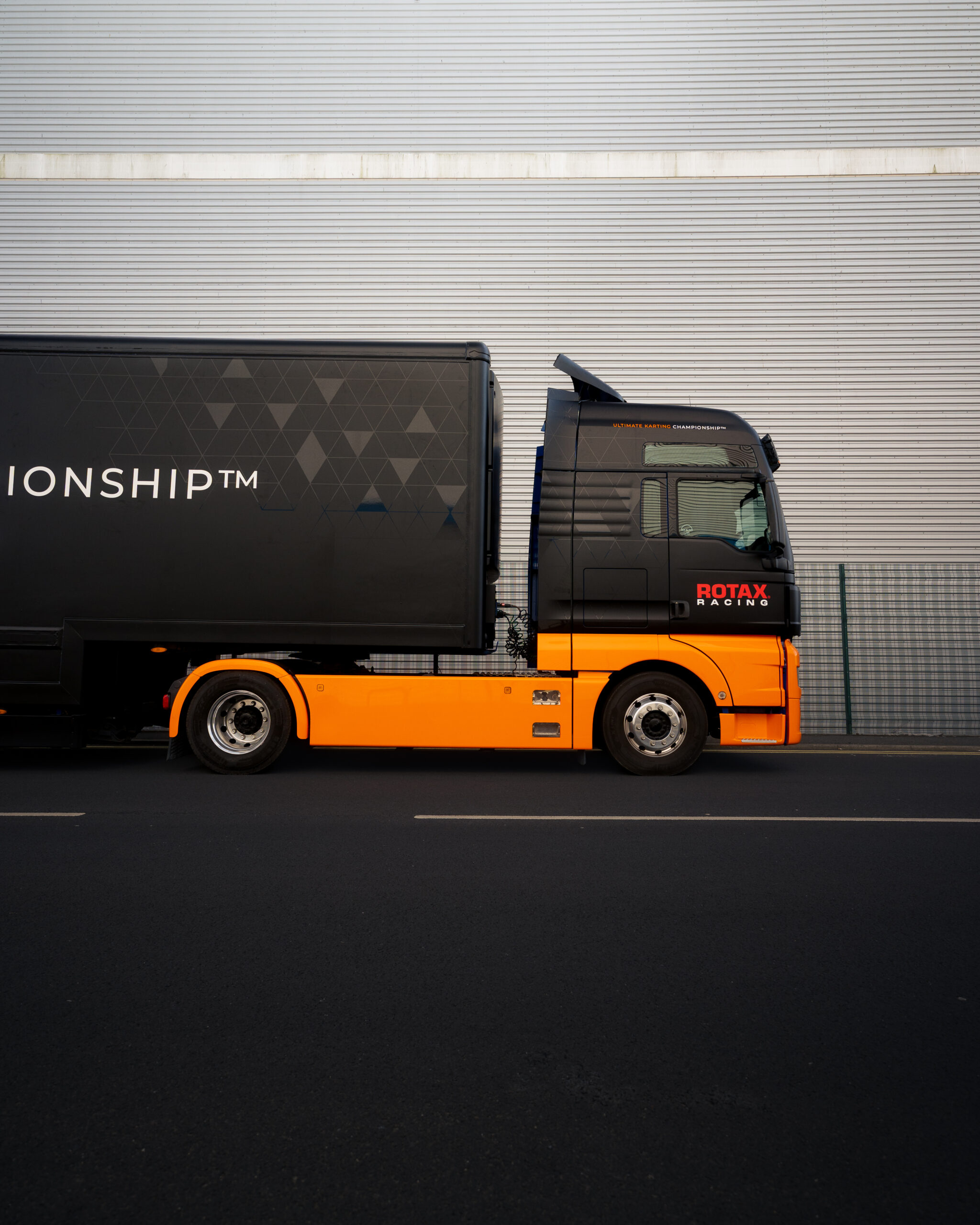 A black and orange semi-truck with "ROTAX" branding is parked on a road in front of a corrugated metal industrial building.