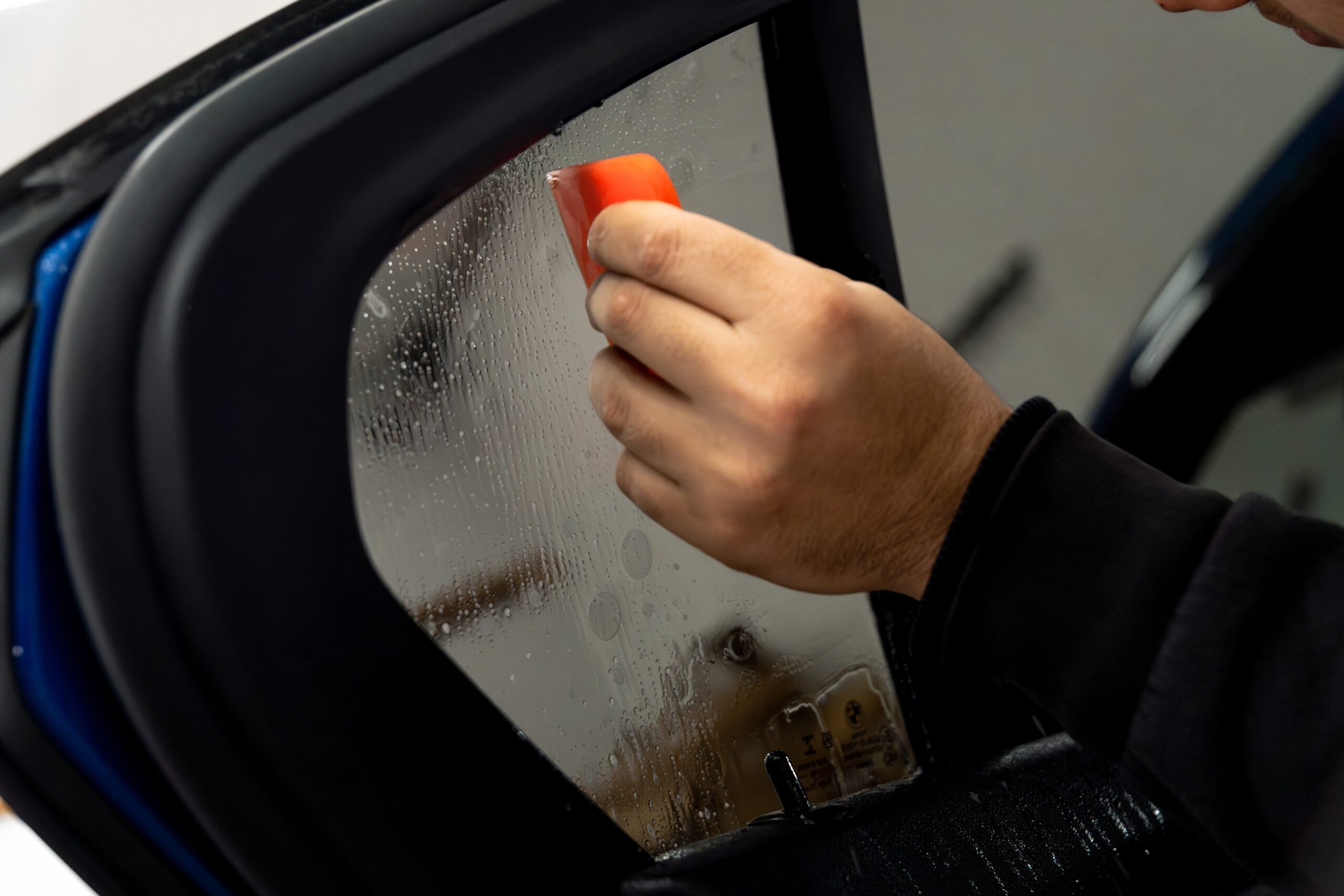 A person uses a small red tool to apply or smooth film onto a car window.