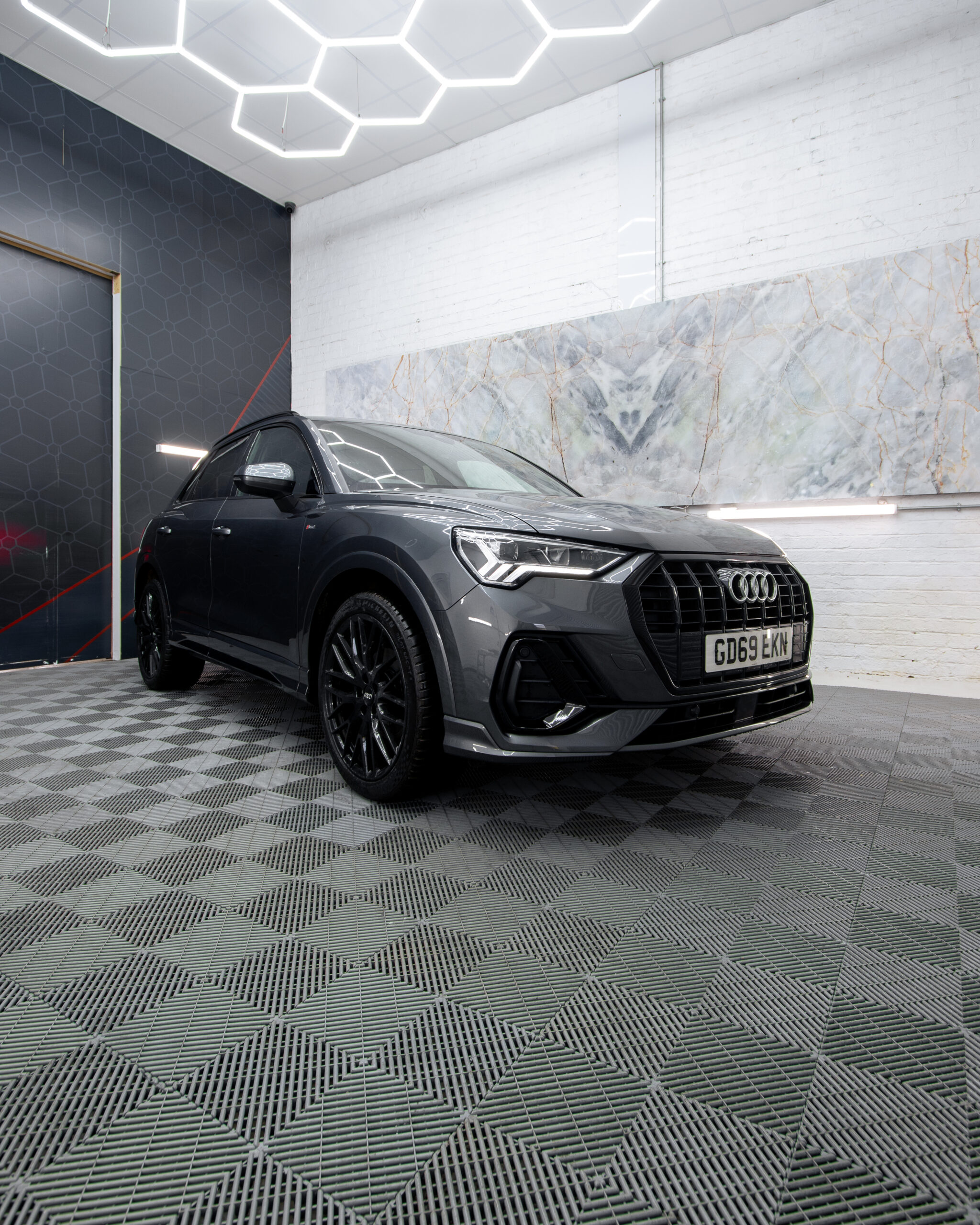A black Audi SUV is parked indoors on a patterned floor, beneath a geometric ceiling light and in front of a marble accent wall.