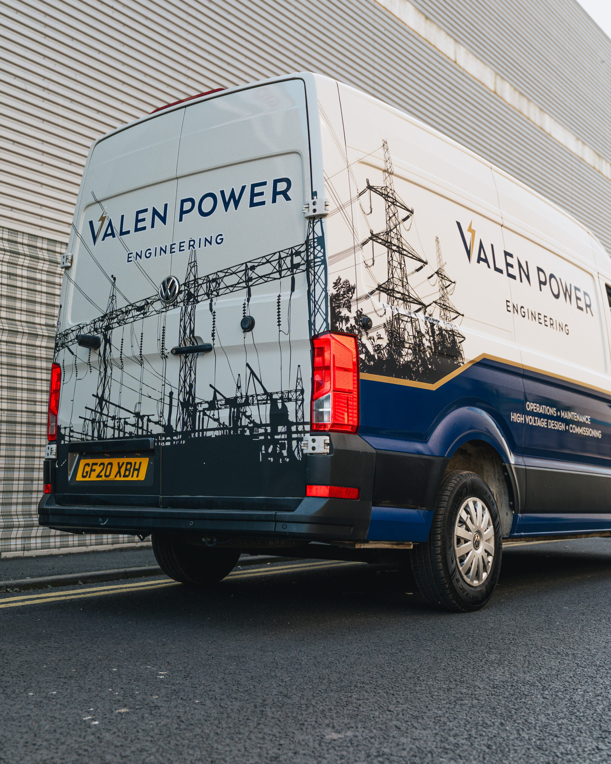 A van with "Valen Power Engineering" branding and power line graphics is parked on a street next to a corrugated metal building.
